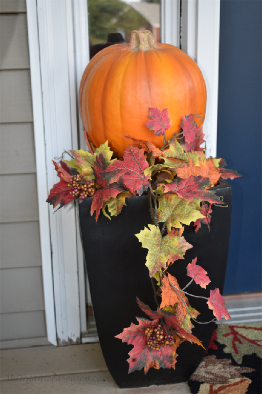 A black planter with an orange pumpkin and a leaf garland on top. Showing how to decorate your front porch for Fall.