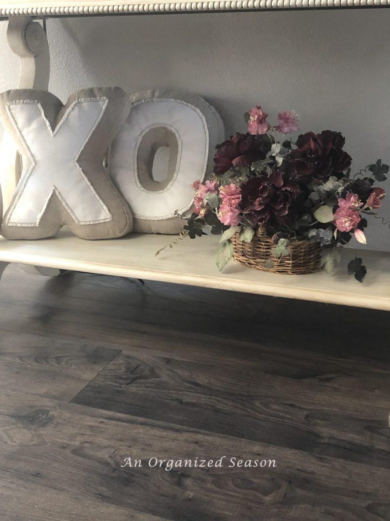 Shelf below foyer table decorated with pillows in the shape of an "X" and an "O" and a flower arrangement