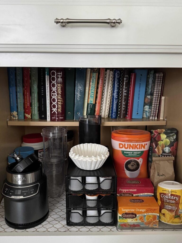 An organized beverage cabinet with coffee pod storage, plastic riser for tea, lemonade, and coffee bags. A smoothie machine, accessories, and protein powders.  Cookbooks line the shelf above. 