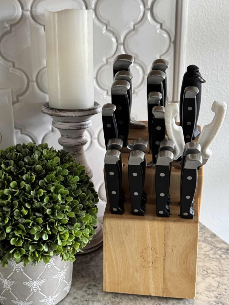 An organized knife set in a wooden block next to a boxwood planter and a pillar candlestick holder.