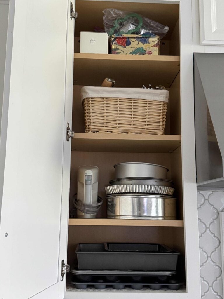 Kitchen cabinet with organized baking pans, mixer, basket, and cookie cutters. 