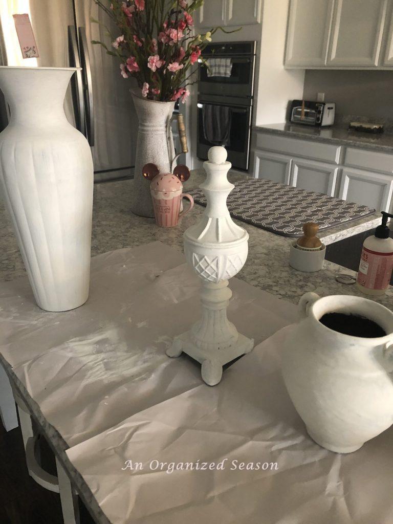 Three old home decor items that have received an update with white paint. There are two vases and a statue sitting on a kitchen counter. 