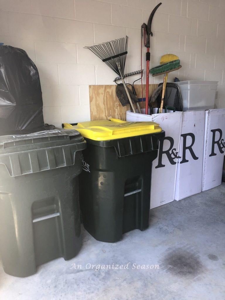 Garage with trash can, recycle, can and three large white cardboard boxes containing lawn tools.  Picture taken before steps to garage organization was started.