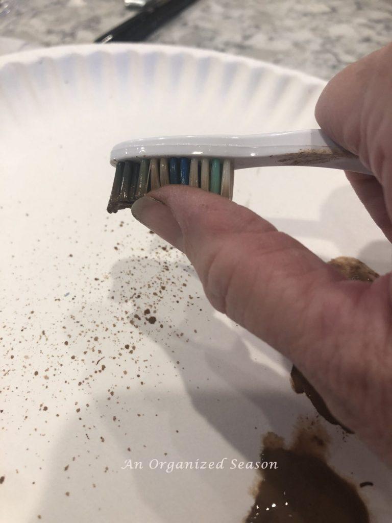 A toothbrush dipped in brown paint being splattered by running a thumb across the bristles.  Showing how to make speckled eggs for Spring decor.