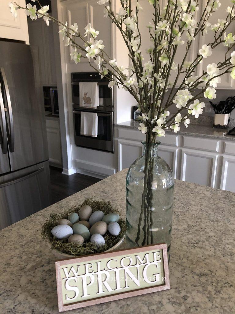 Speckled eggs displayed on a white cake plate with a "welcome spring" sign and a vase of flowers.  An example of how to make speckled eggs and then to dispaly them in Spring decor. 