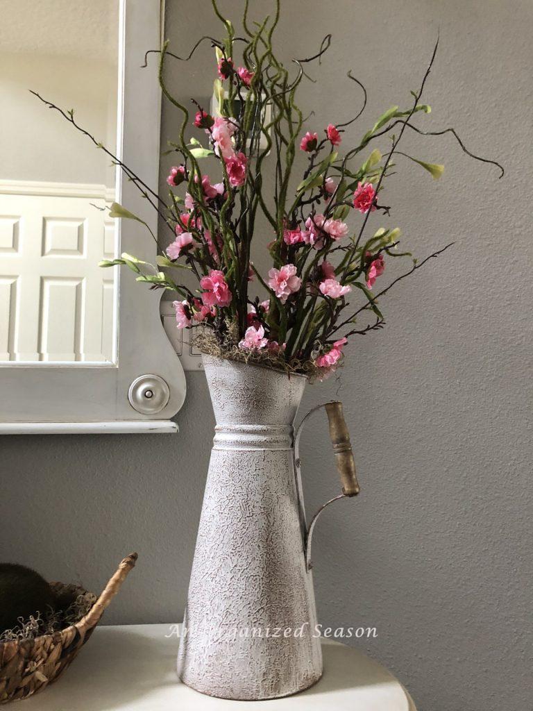 Bright pink flowers on tree branches inside a metal pitcher painted white. An example of helpful ideas to brighten up spring decor.