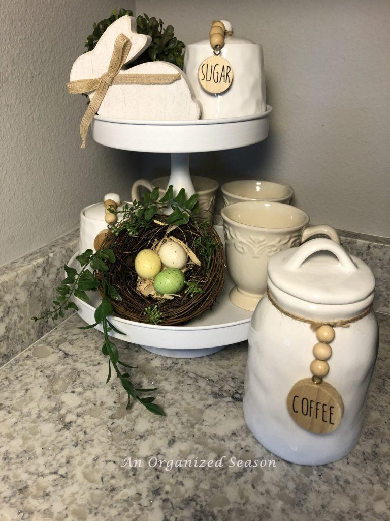 White two tiered tray holding three coffee cups, two lidded containers, a nest with bird eggs, a wooden bunny.  A white container holding coffee is beside the tray on a kitchen counter.  Example of helpful ideas to brighten the spring decor in your home. 