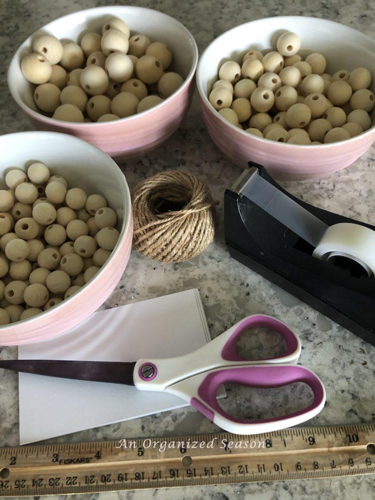 Three bowls with different size wooden beads, a roll of jute twine, scotch tape dispenser, scissors and index cards, and a ruler. Items needed to complete how to make a wood bead garland with tassels.