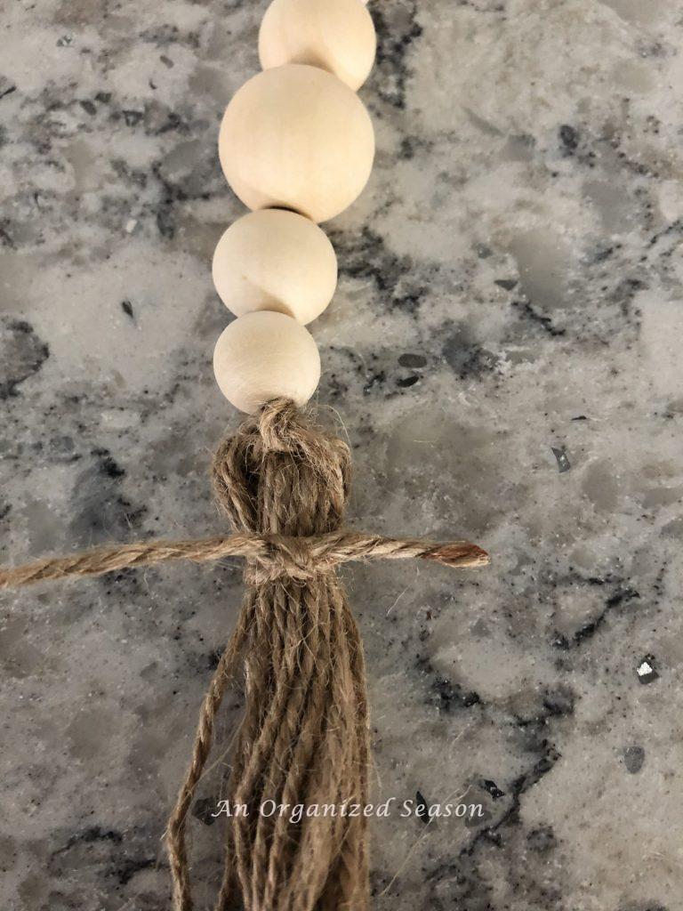 Showing how to tie a knot to hold a jute twine tassel together. Second to last step in how to make a wood bead garland with tassels. 