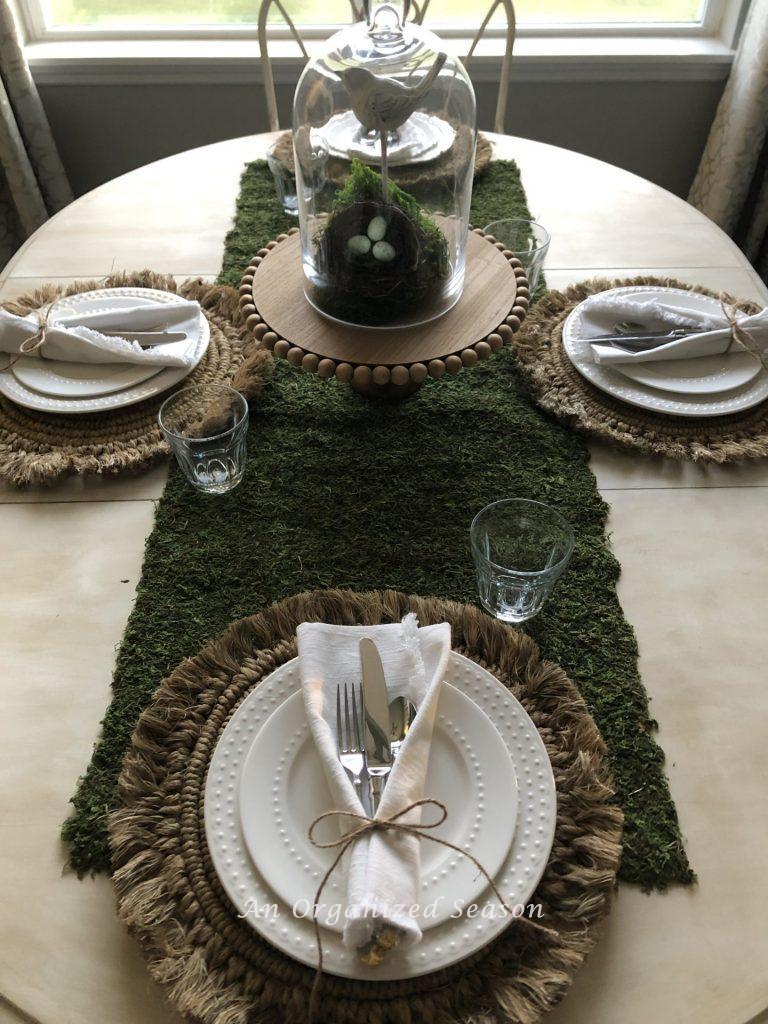 A white kitchen table set with a moss runner, four white place settings and a centerpiece showing helpful ideas for how to brighten your spring decor. 