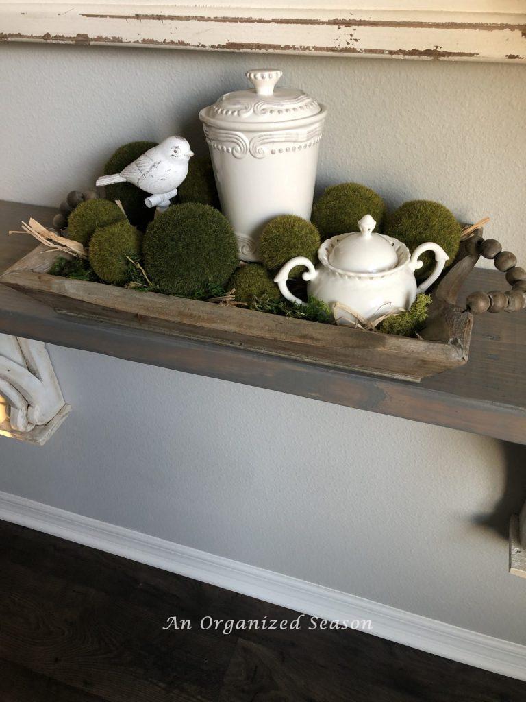 A skinny wooden bowl holding two white containers with lids and a white bird statue, surrounded by bright green moss balls of varying sizes.  One of several helpful ideas for how to brighten up your Spring decor. 