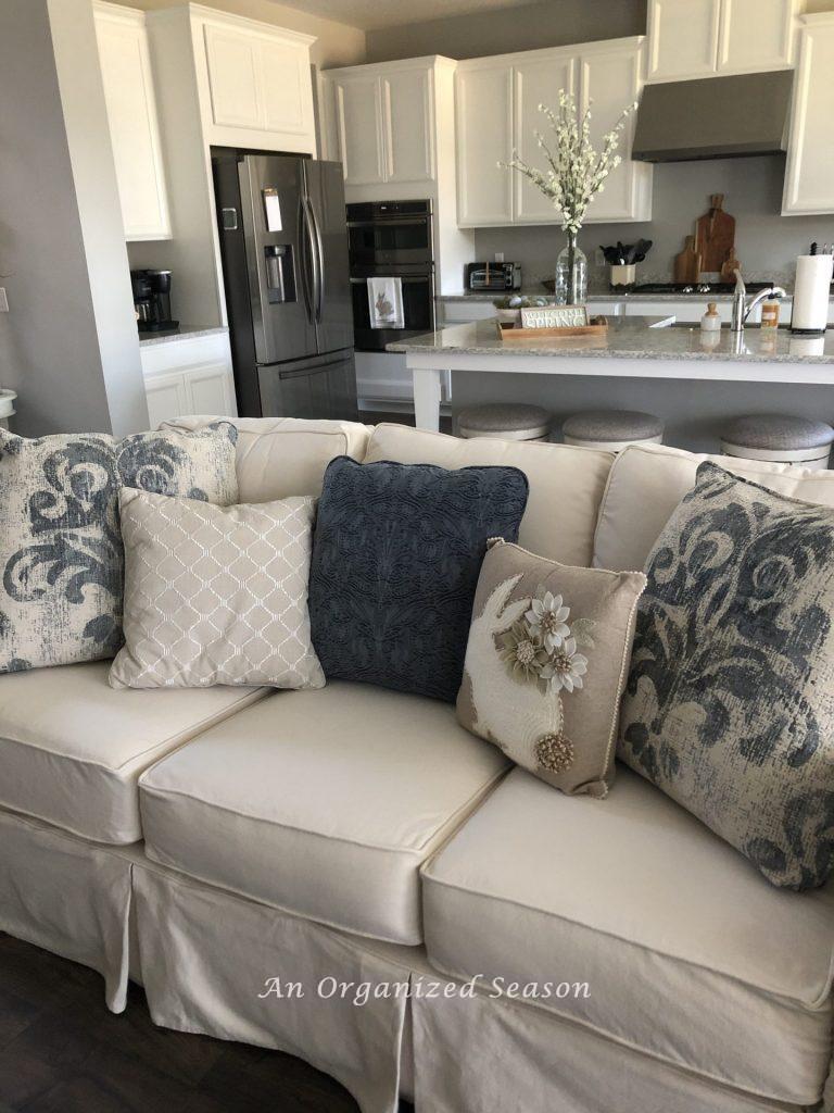 A white slip-covered couch with two large blue and white pillows, a medium size blue denim pillow, a small taupe pillow with white embroidery lines and a small taupe pillow with a white bunny and felt flowers.  All helpful ideas to brighten up spring your decor. 