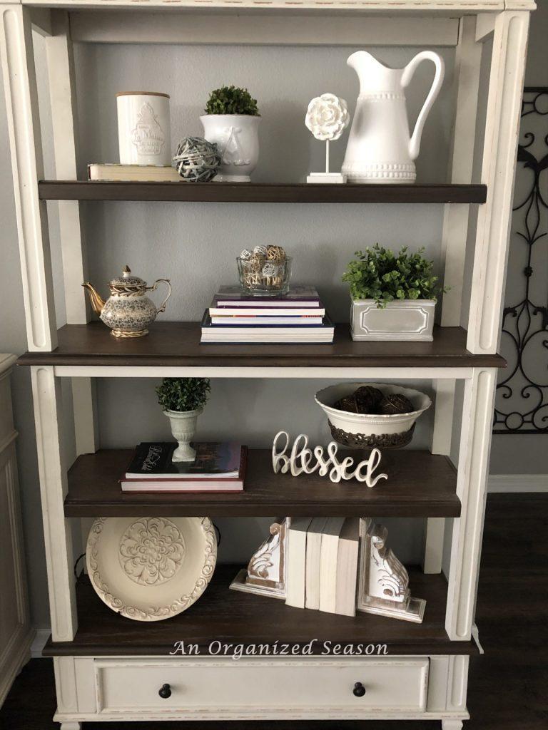 A bookshelf styled with white dishes, stacks of books turned backwards and three potted plants showing helpful ideas to brighten your spring decor.