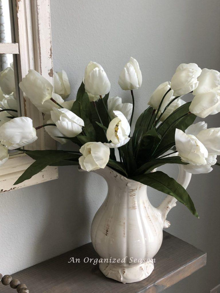 White tulips in a white pitcher sitting on a shelf.  one of many  helpful ideas for how to brighten your Spring decor.