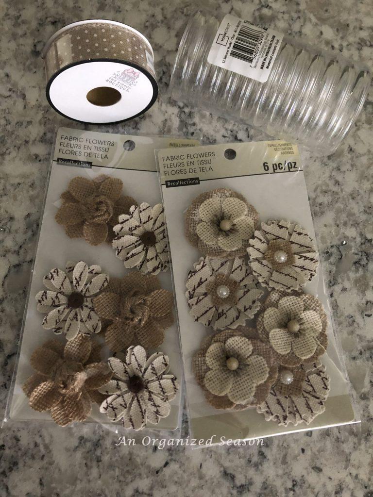 Two packages of fabric flowers, a roll of ribbon, and a package of clear plastic shower curtain holders laying on kitchen counter.  Items needed to create inexpensive floral napkin rings.