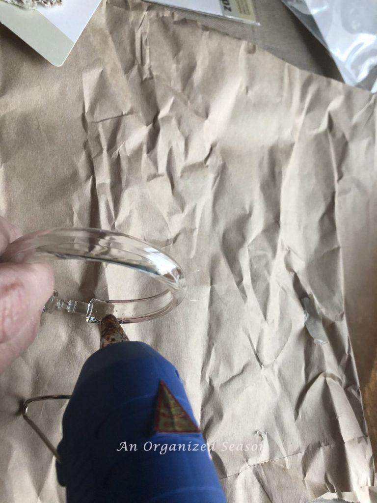 A glue gun squirting glue on a clear shower curtain ring showing step one of how to create an inexpensive floral napkin ring.