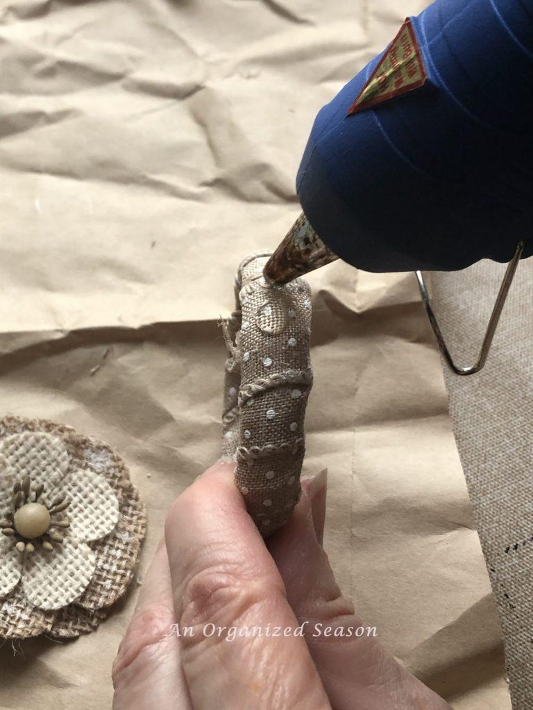 A hot glue gun squirting glue on the top of a shower curtain ring wrapped with ribbon.  Showing step six of how to create an inexpensive floral napkin ring.