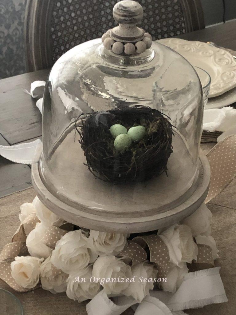 A white wooden cake plate holding a bird nest with three small blue eggs that is covered by a glass cloche. An example of an easy centerpiece giving tips for creating a simple Easter tablescape. 