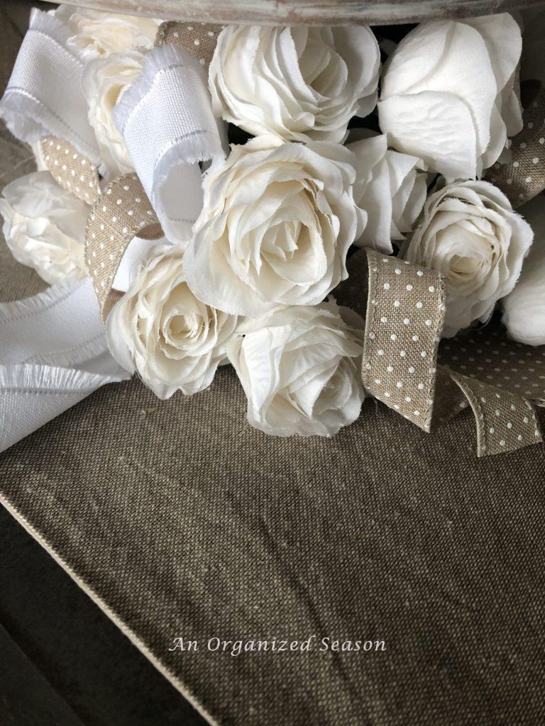 White roses surrounding the base of a white wooden cake plate with white and beige polka dotted ribbon running  between the flowers.  An example of tips to create a simple Easter tablecape.