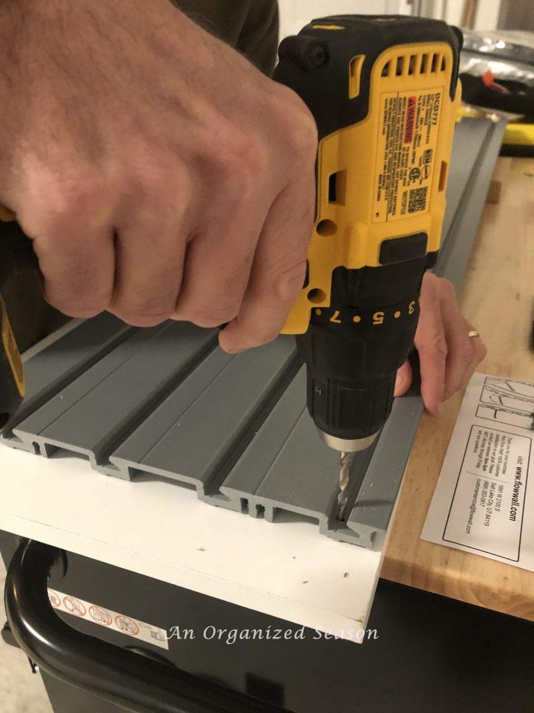 A man's hand holding a drill on a Flow Wall panel.  Step two for how to install a Flow Wall system. 