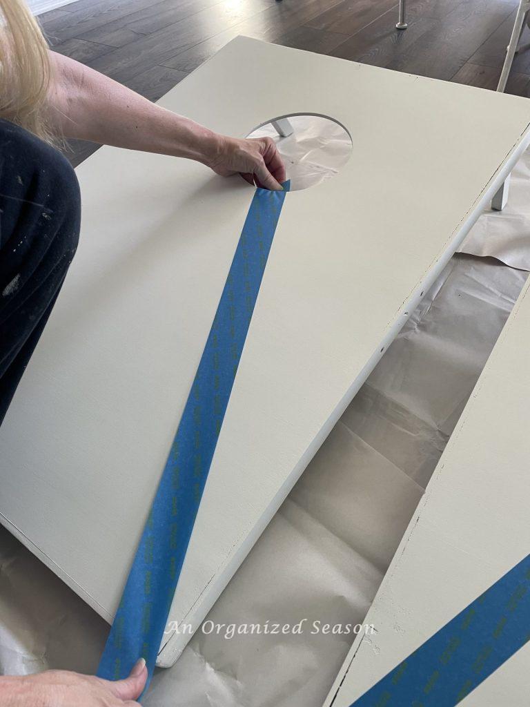 A woman placing painter's tape on a cornhole board.  Showing step six for steps to transform a worn out cornhole set.