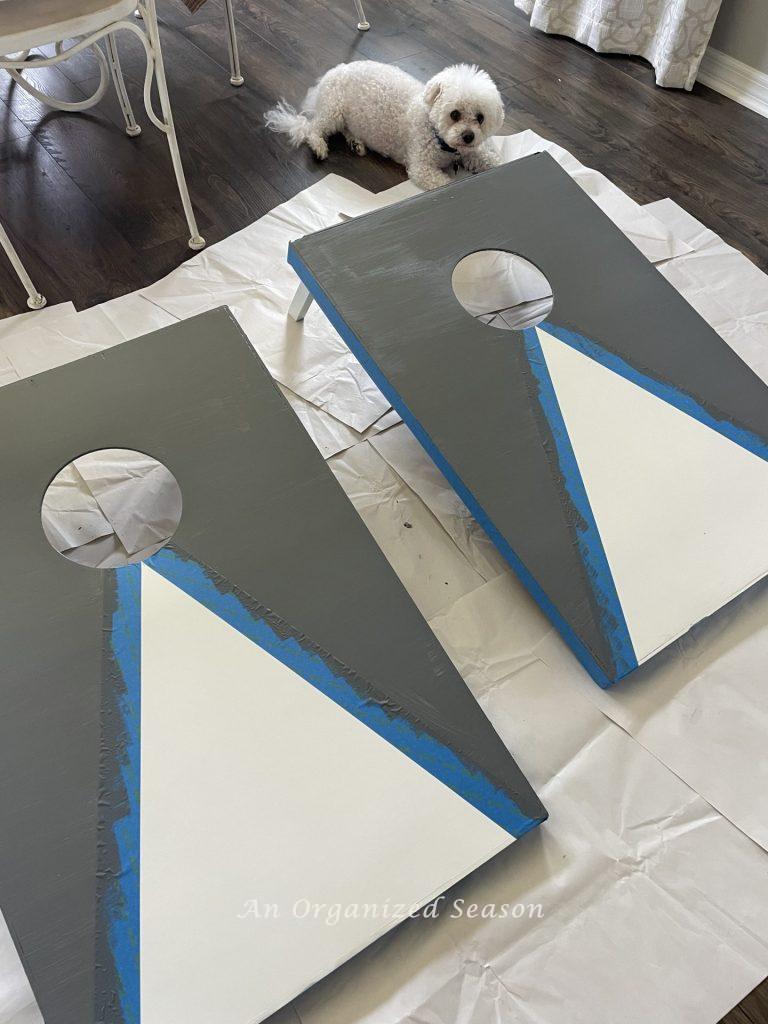 Two cornhole boards being painted with gray and white paint. Showing step seven for steps to transform a worn out cornhole set.