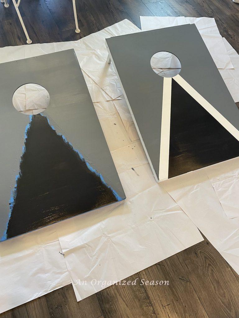 A cornhole board with the painters tape removed showing white lines. Another board beside it with painter's tape still on. Step nine for steps to transform a worn out cornhole set.