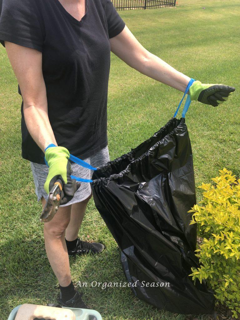 A woman in a yard with a black garbage bag.  Her wrists are inside the handles of the bag, holding it open. Step one for helpful tips to organize and spruce up your yard.