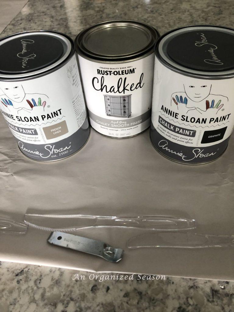 Three cans of paint sitting on paper on a kitchen counter with a paint can opener and a plastic knife to stir the paint. Showing step five for how to makeover a breakfast in bed tray.