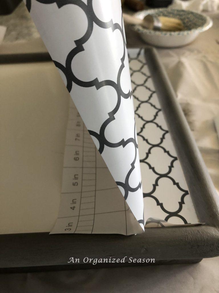 A gray bed tray that is being covered with contact paper on the top surface.  Approximately two inches of the contact paper has been applied.  Showing step nine for how to makeover a breakfast in bed tray. 