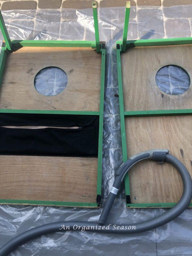 A cornhole set turned upside down with a vacuum cleaner laying beside it.  Showing step one of for steps to transform a worn out cornhole set.