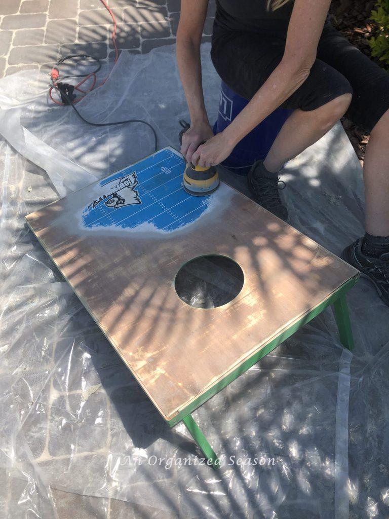 A woman sitting next to a cornhole board sanding the old paint off the top.  Shows step two for steps to transform a worn out cornhole set. 