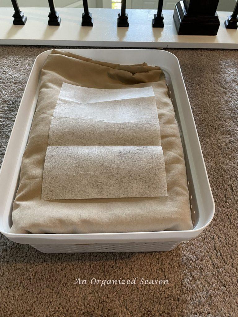 A sheet set folded and placed inside a white storage container with a dryer sheet laying on top. An idea of how to organize linen and cleaning closets.