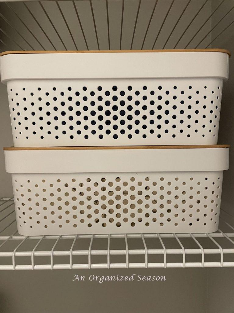 Two white containers with holes and bamboo lids stacked on top of each other on a wire shelf.  An idea on how to organize linen and cleaning closets.
