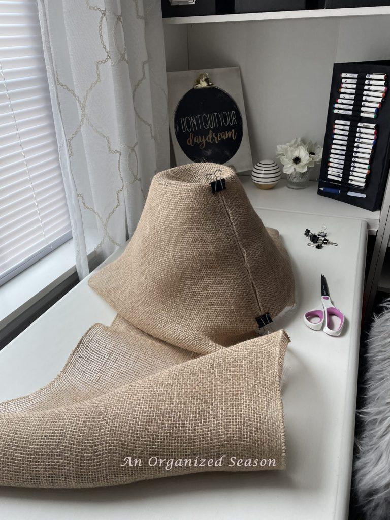 A lamp shade wrapped in burlap, sitting on a desk next to a pair of scissors.  Step one for how to makeover an old lamp to match new decor.