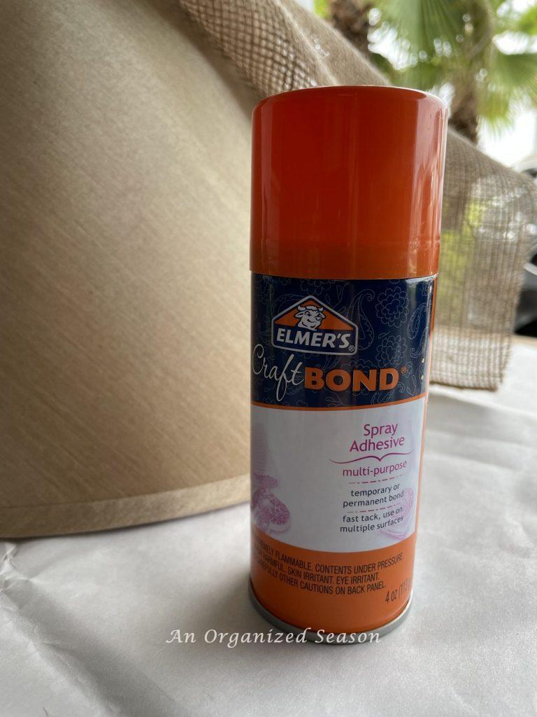 A can of Elmer's Craft Bond spray adhesive sitting in front of a lampshade. Showing a product to use to makeover an old lamp to match new decor. 