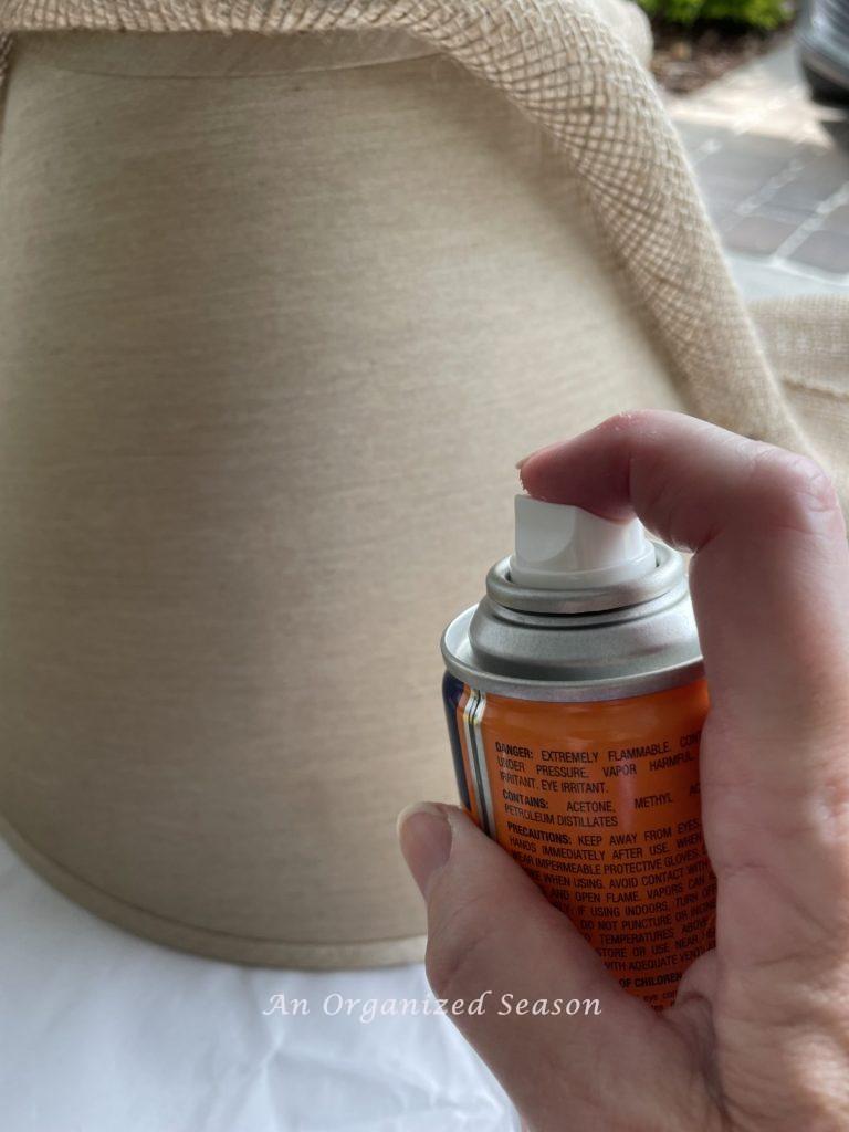 A hand spraying adhesive on a lamp shade. Showing step six for how to makeover an old lamp to match new decor.