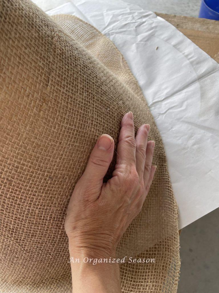 A hand applying burlap to an old lampshade. Step five for how to makeover an old lamp to match new decor. 