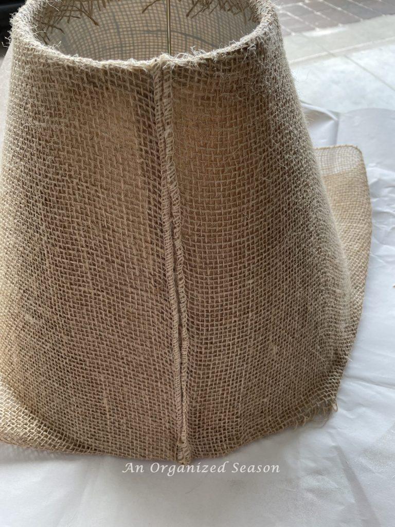 A lamp covered in burlap with a seam that was hot glued down.  Showing step seven for how to makeover an old lamp to match new decor.