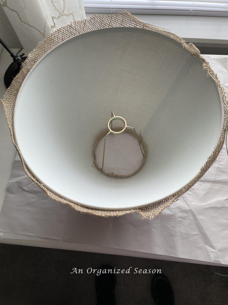 A lamp shade turned upside down with a one inch edge of burlap sticking out. Step eight for how to makeover an old lamp to match new decor.