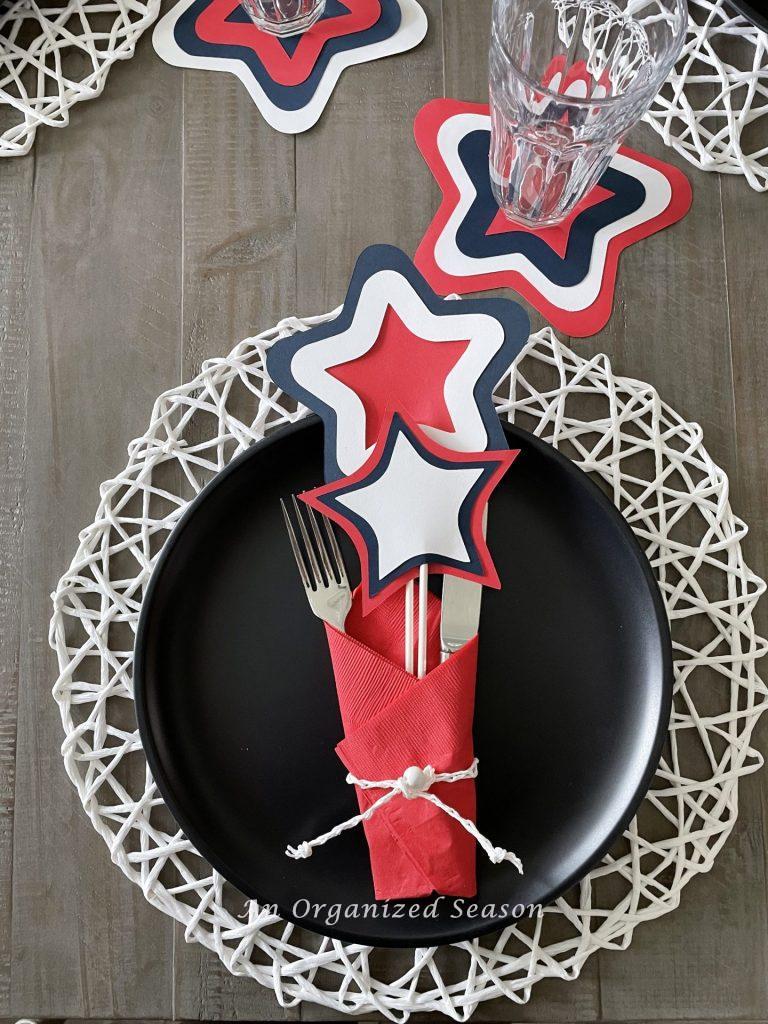 A place setting on a dining room table consisting of a white placemat, black dinner plate, silver fork and knife wrapped in a red napkin that's been tied with a white cord. The are two paper red, white and blue stars tucked inside the napkin, showing how to create a patriotic table for the 4th of July!