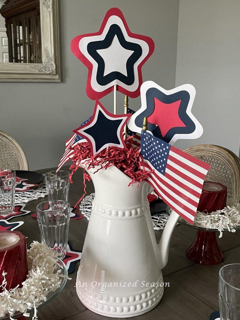 A white pitcher with 3 small US flags and 3 red, white, and blue stars made from paper and glued on white sticks, showing how to create a patriotic table for the 4th of July.