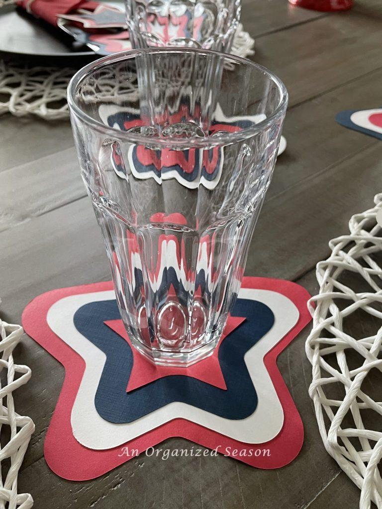 A clear drinking glass sitting on a coaster made out of three red, white, and blue stars showing an example of how to create a patriotic table for the 4th of July. 