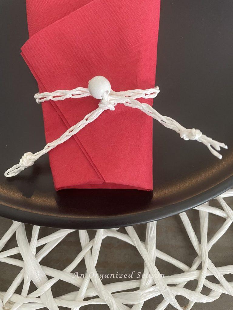 A red napkin that has been folded around eating utensils then wrapped in a white cord with a white bead, which is an example of how to create a patriotic table for the 4th of July.