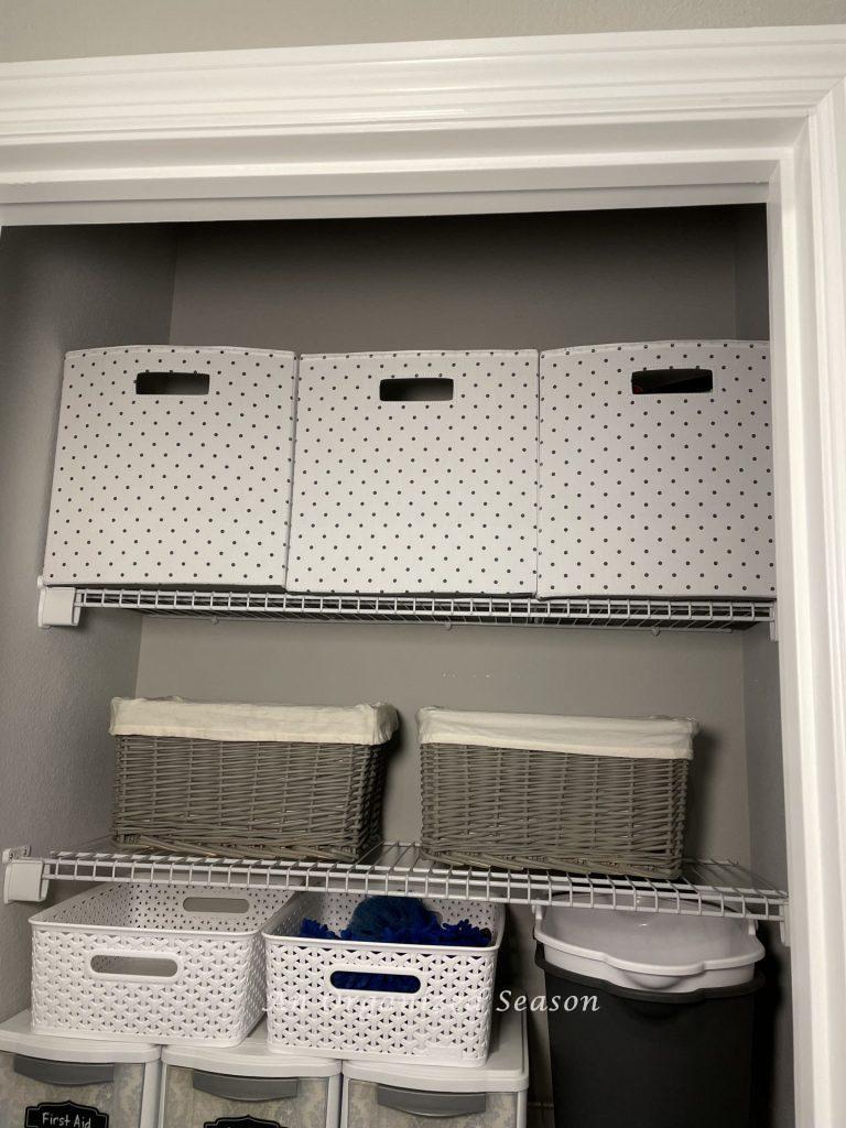 Three polka dotted storage cubes and two gray baskets sitting on wire shelves in a cleaning closet. Showing ideas to organize linen and cleaning closets. 