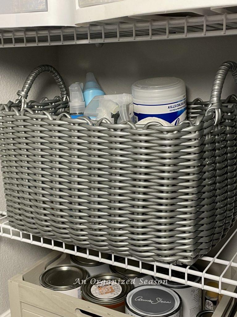 A gray plastic basket with handles containing cleaning products, sitting on a wire shelf in a cleaning closet. An idea for how to organize linen and cleaning closets. 
