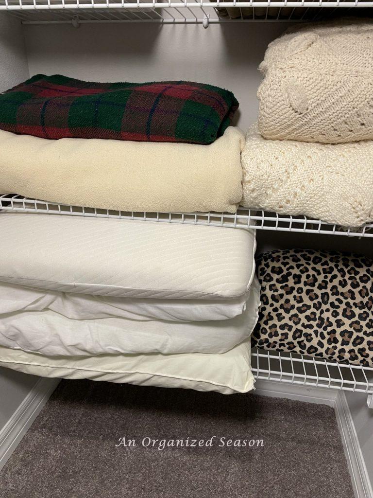 Two wire shelves in a closet holding a stack of pillows and two stacks of blankets. An idea of how to organize linen and cleaning closets. 