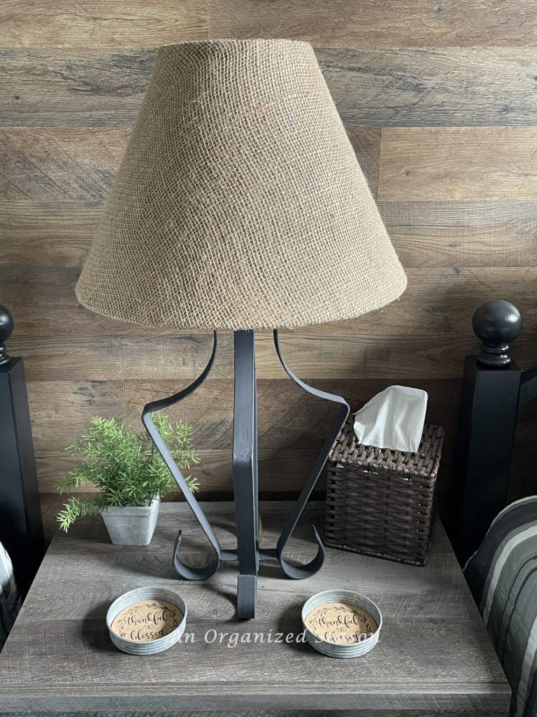 A rustic table lamp sitting on a bedside table with weaved tissue holder, plant, and rustic cup holders. Showing step seven for how to makeover a bedroom into a rustic retreat. 