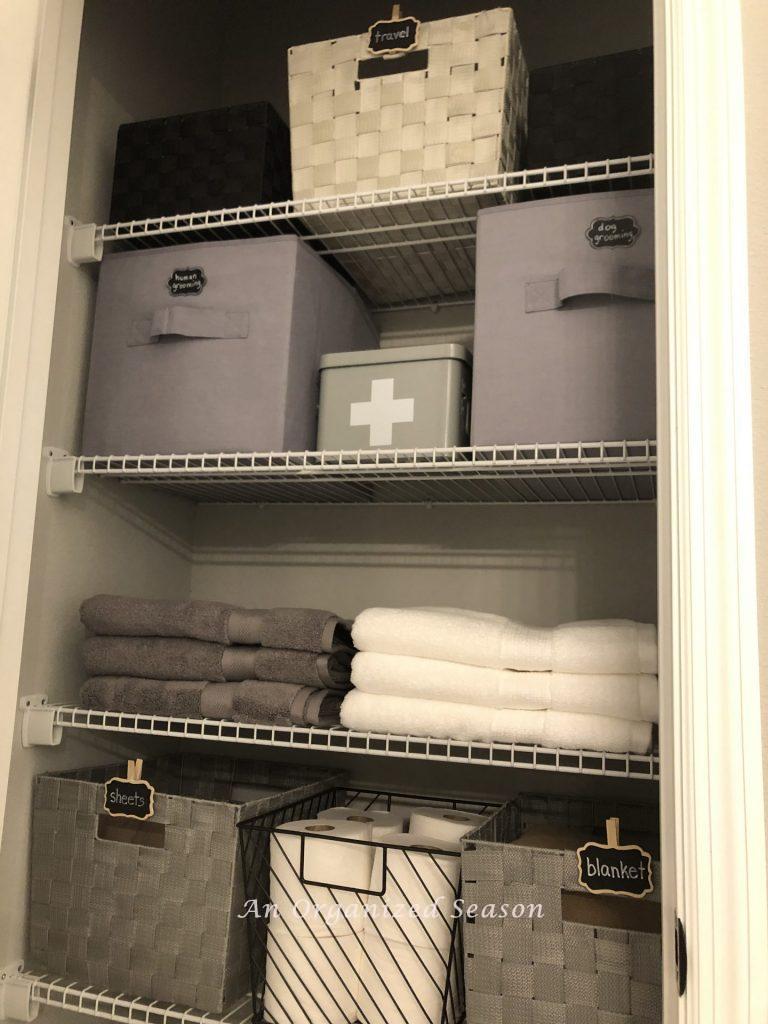 An organized bathroom closet with gray bins, towels, a first aid kit, and a toilet paper holder. The result of how to make home organization goals.