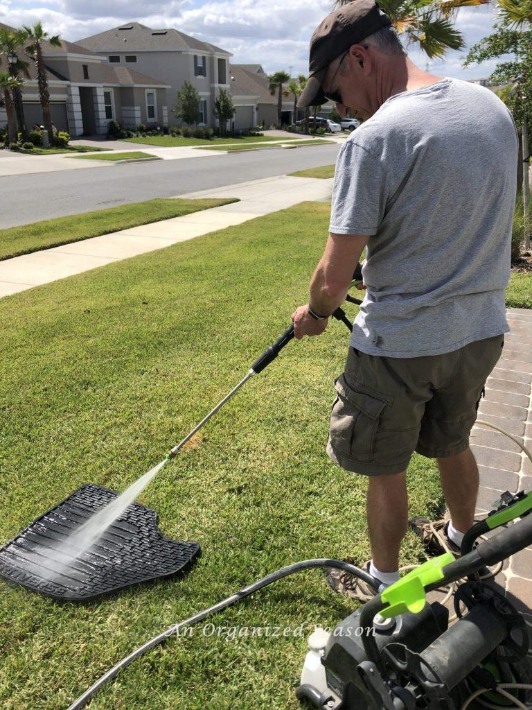 A man using a power washer to clean a car floormat in his front yard. A tip for how to organize and clean your car.